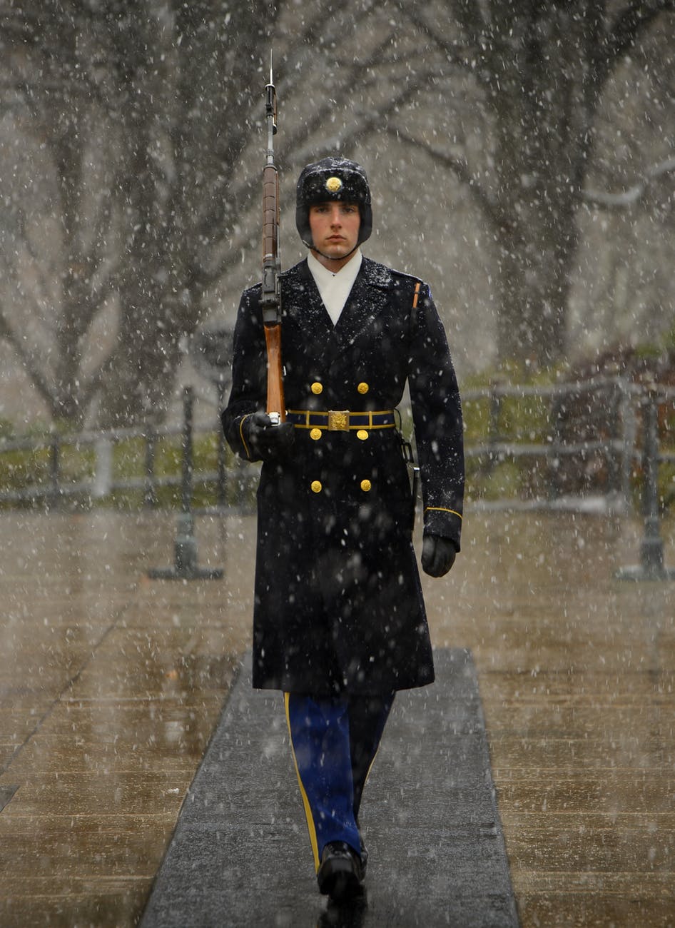 man in black coat carrying a rifle walking in straight line during daytime